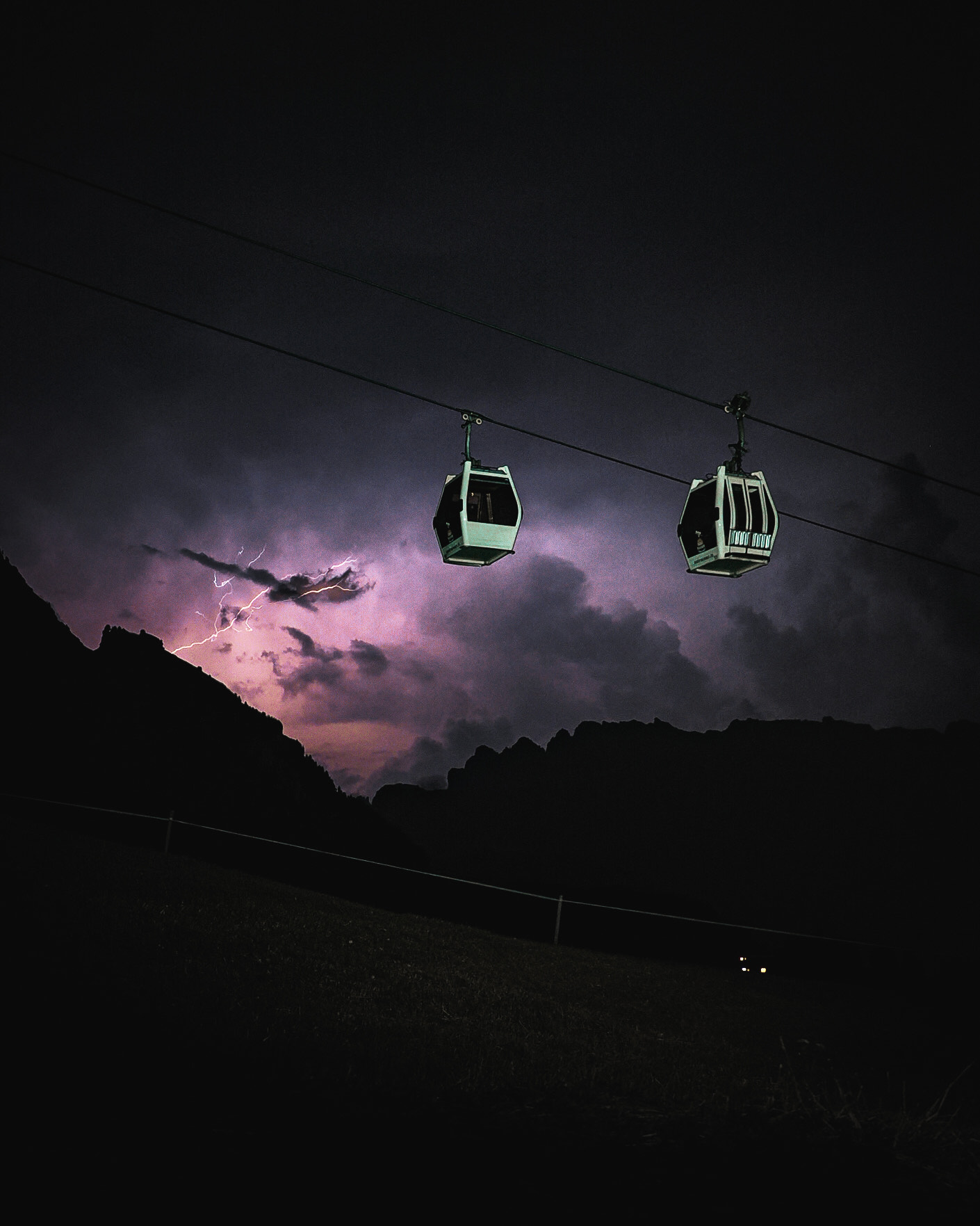 A photo of dark sky and hills with lightning in the sky, with cablecars lit up by torchlight in the foreground