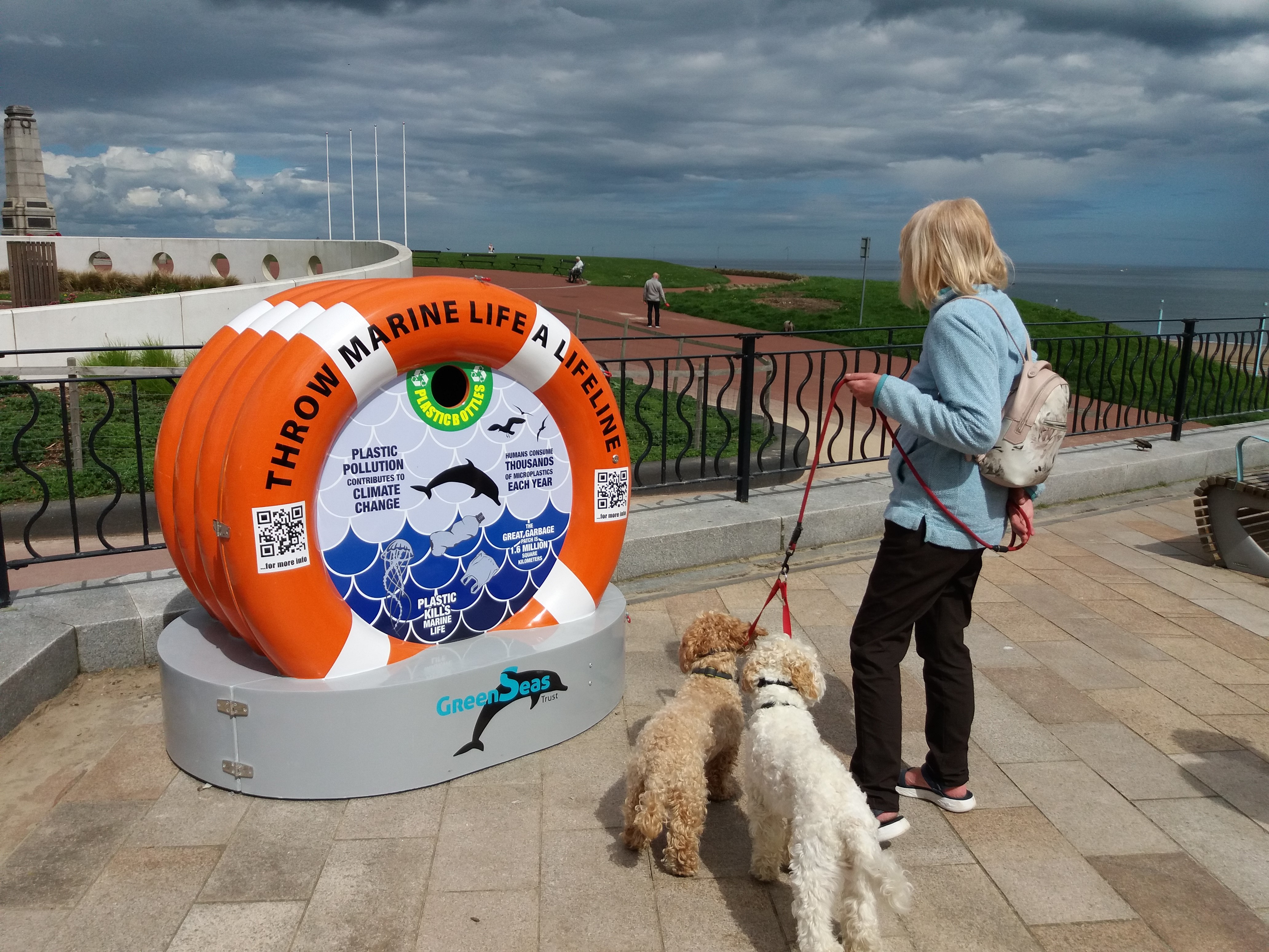 A woman and her two dogs stand facing a orange buoy-shaped bin on a seaside promenade.