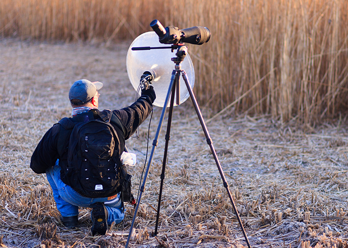 A person wearing a cap and backpack kneels outside next to a camera on a tripod, holding up a large microphone next to some long grass.