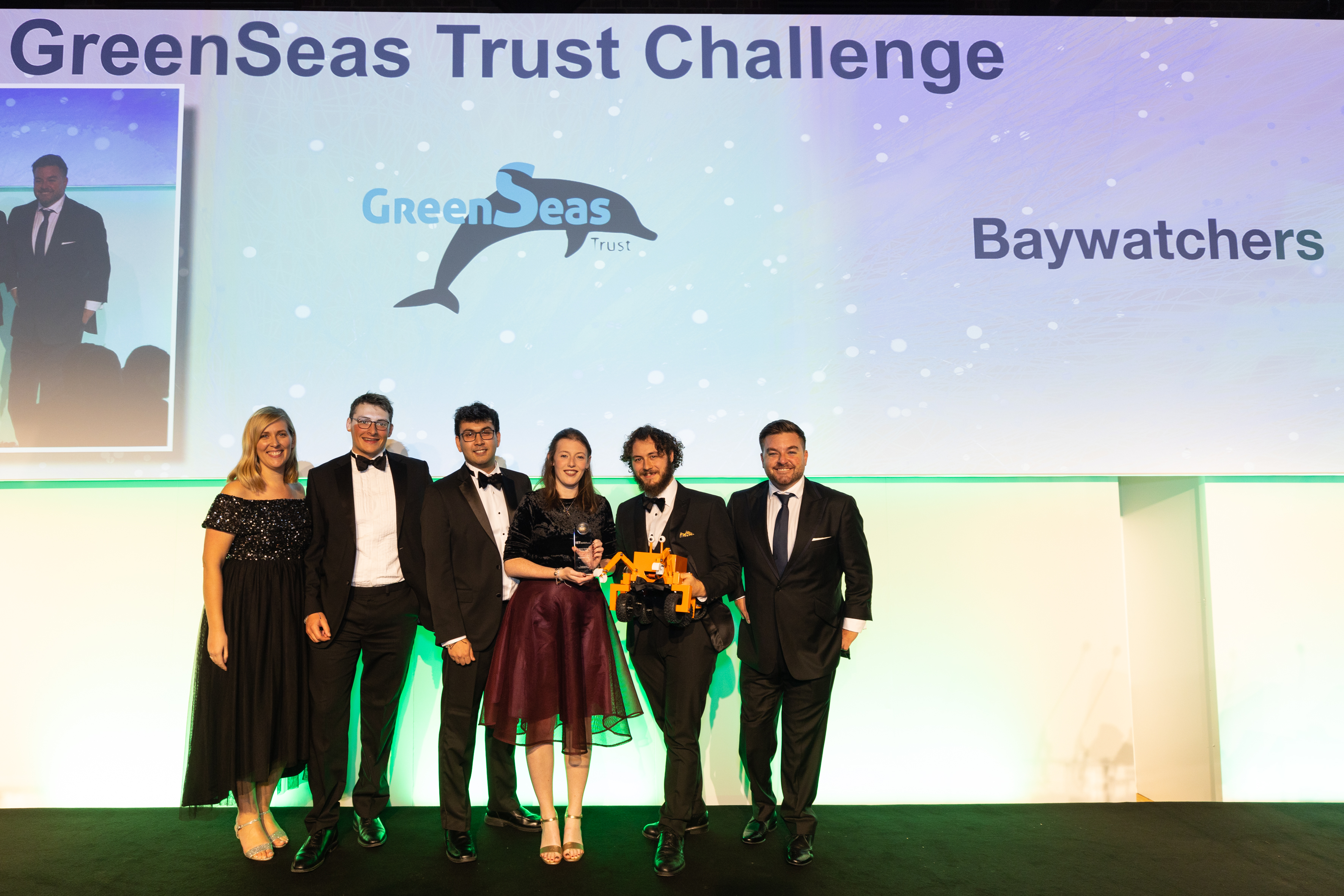 A photo of 6 people standing on a stage in formal dress, in front of a screen which reads GreenSeas Trust Challenge: Baywatchers.