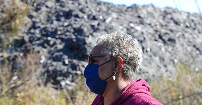 Marsha Jackson wears a face mask, and stands side on to the camera, in front of a large grey mountain of roof shingles in the background.