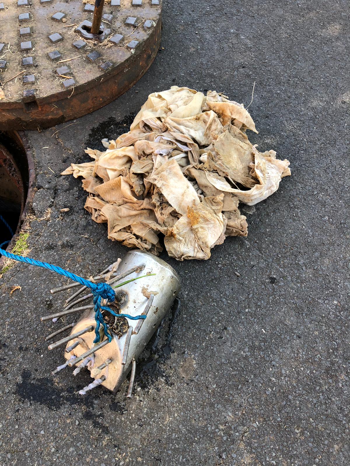 A photo of a cylindrical metal tool with a series of spikes, next to a pile of discoloured wet wipes and an open sewer grate.