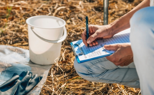 Person crouching and recording notes about soil. They have a bucket and gloves nearby.