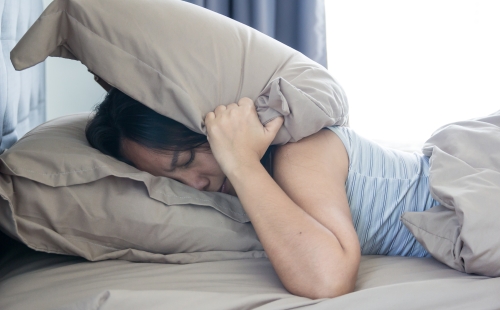 Young woman lying in bed suffering from sound covering head and ears with pillow making unpleasant face