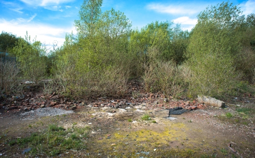 Abandoned site of former industrial gas works. There is stony rubble on the ground with vegetation surrounding it.