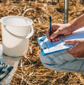Person crouching and recording notes about soil. They have a bucket and gloves nearby.